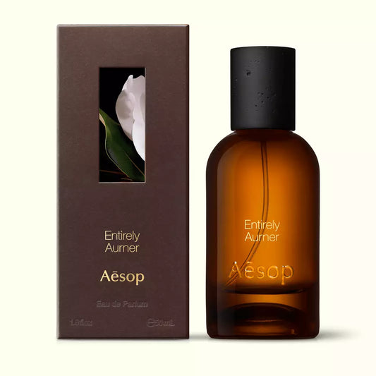 Amber glass Aesop Entirely Aurner Eau de Parfum bottle beside its brown outer box on a white background.