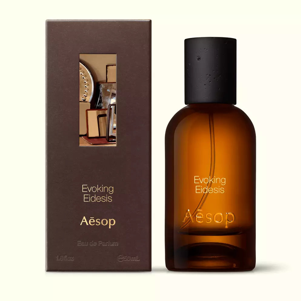 Amber glass Aesop Evoking Eidesis Eau de Parfum bottle beside its brown outer box on a white background.