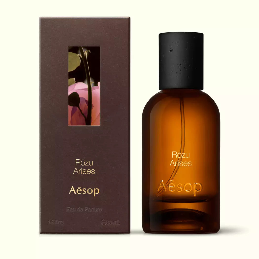 Amber glass Aesop Rozu Arises Eau de Parfum bottle beside its brown outer box on a white background.
