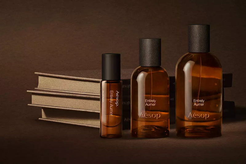 Three amber Aesop fragrance bottles arranged beside stacked cream books on a dark brown surface.