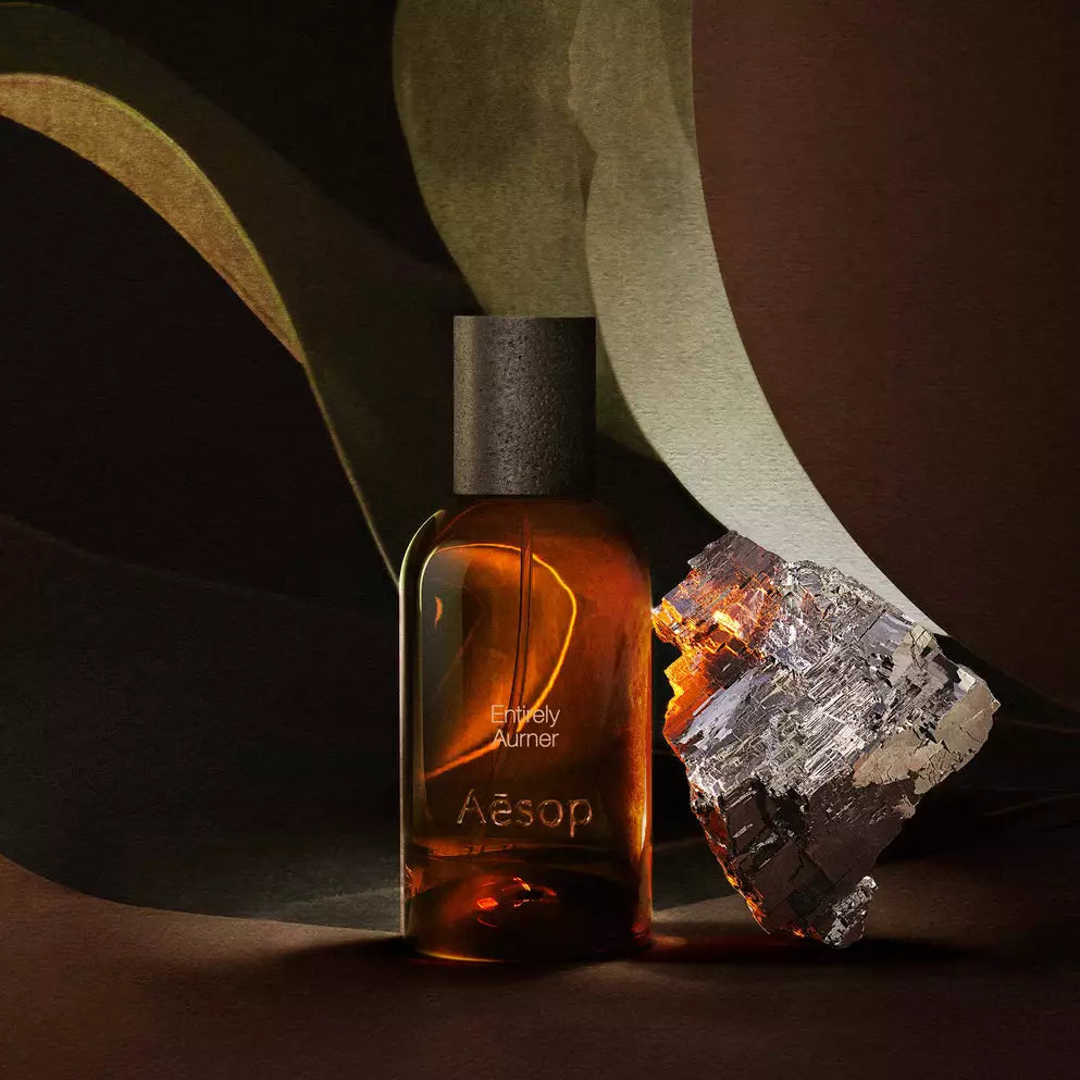 Aesop Entirely Aurner bottle beside a mineral fragment and broad green leaves.