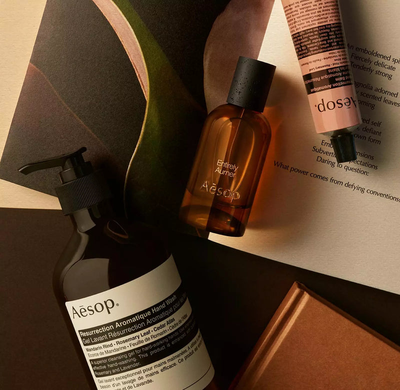 Aesop Entirely Aurner bottle with pump bottle and peach tube arranged on papers and curved dark shapes.