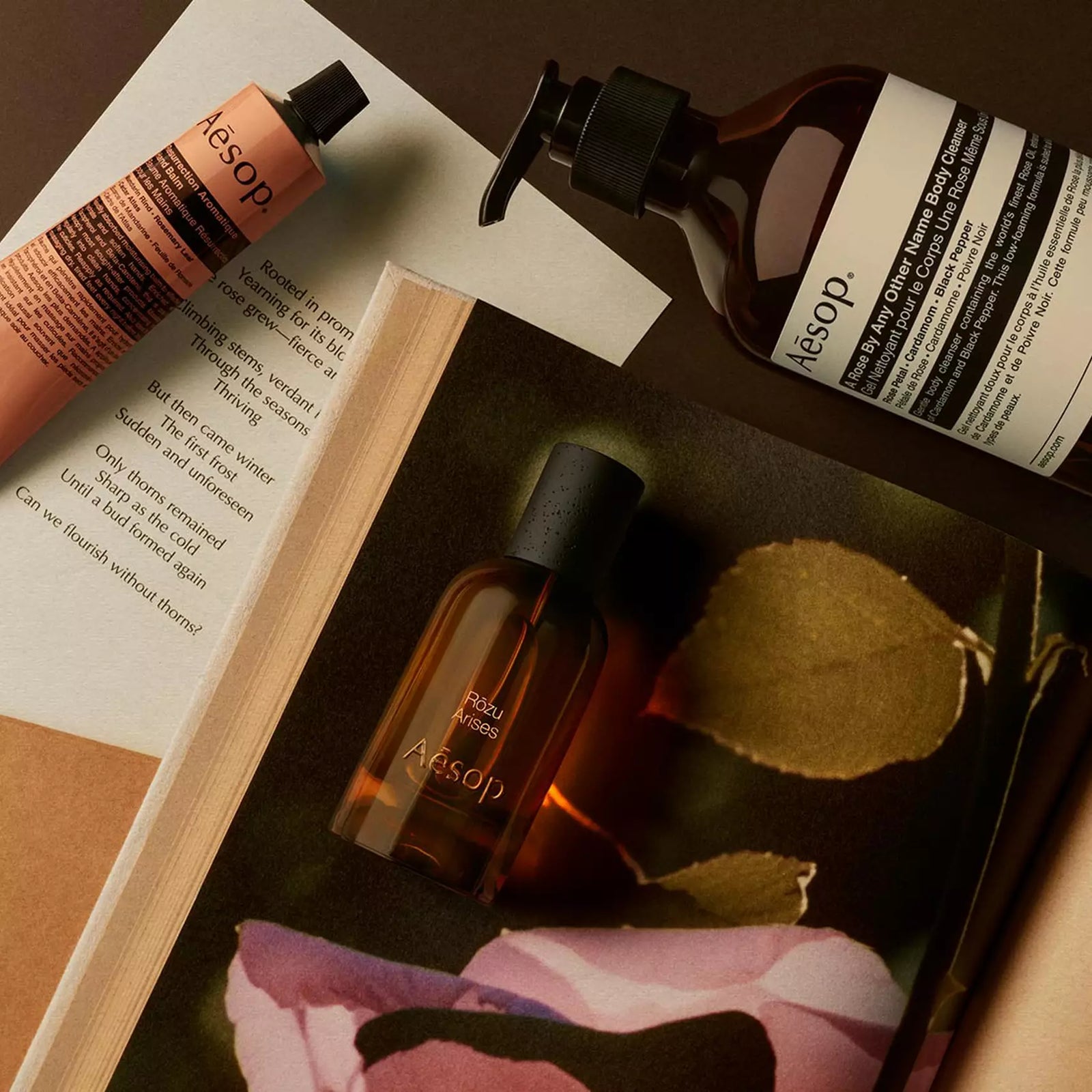 Aesop Rozu Arises bottle with pump bottle and peach tube arranged on papers with pink petals and a leaf.