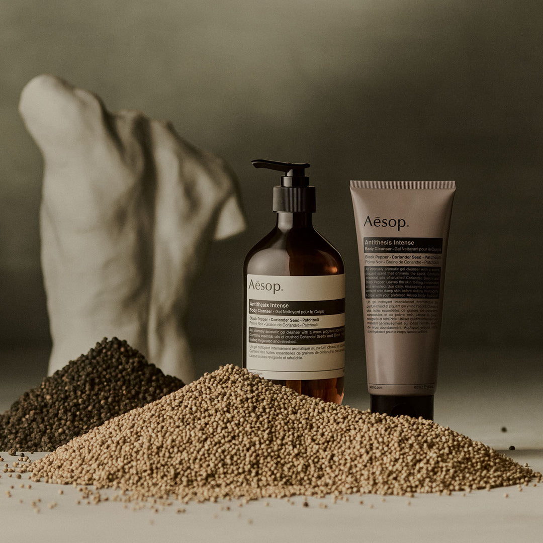 Aesop Antithesis Intense Body Cleanser pump bottle and tube beside piles of black peppercorns and coriander seeds.