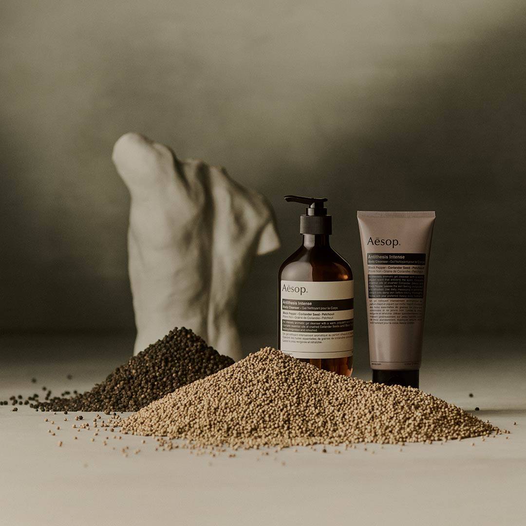 Aesop Antithesis Intense Body Cleanser pump bottle and tube beside piles of black peppercorns and coriander seeds.