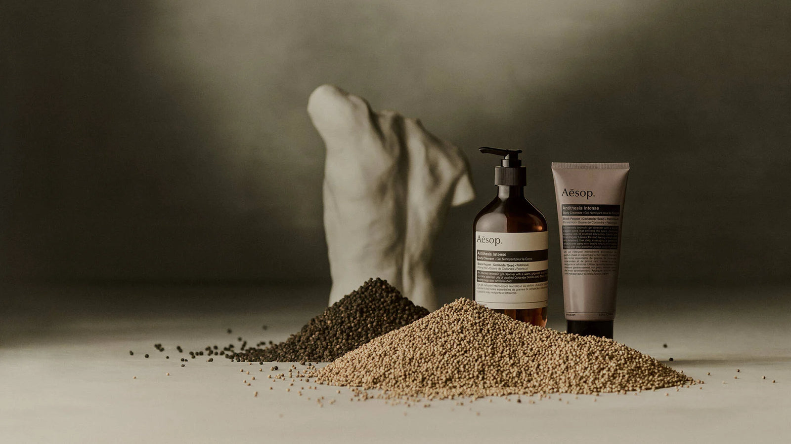 Aesop Antithesis Intense Body Cleanser pump bottle and tube beside piles of black peppercorns and coriander seeds.